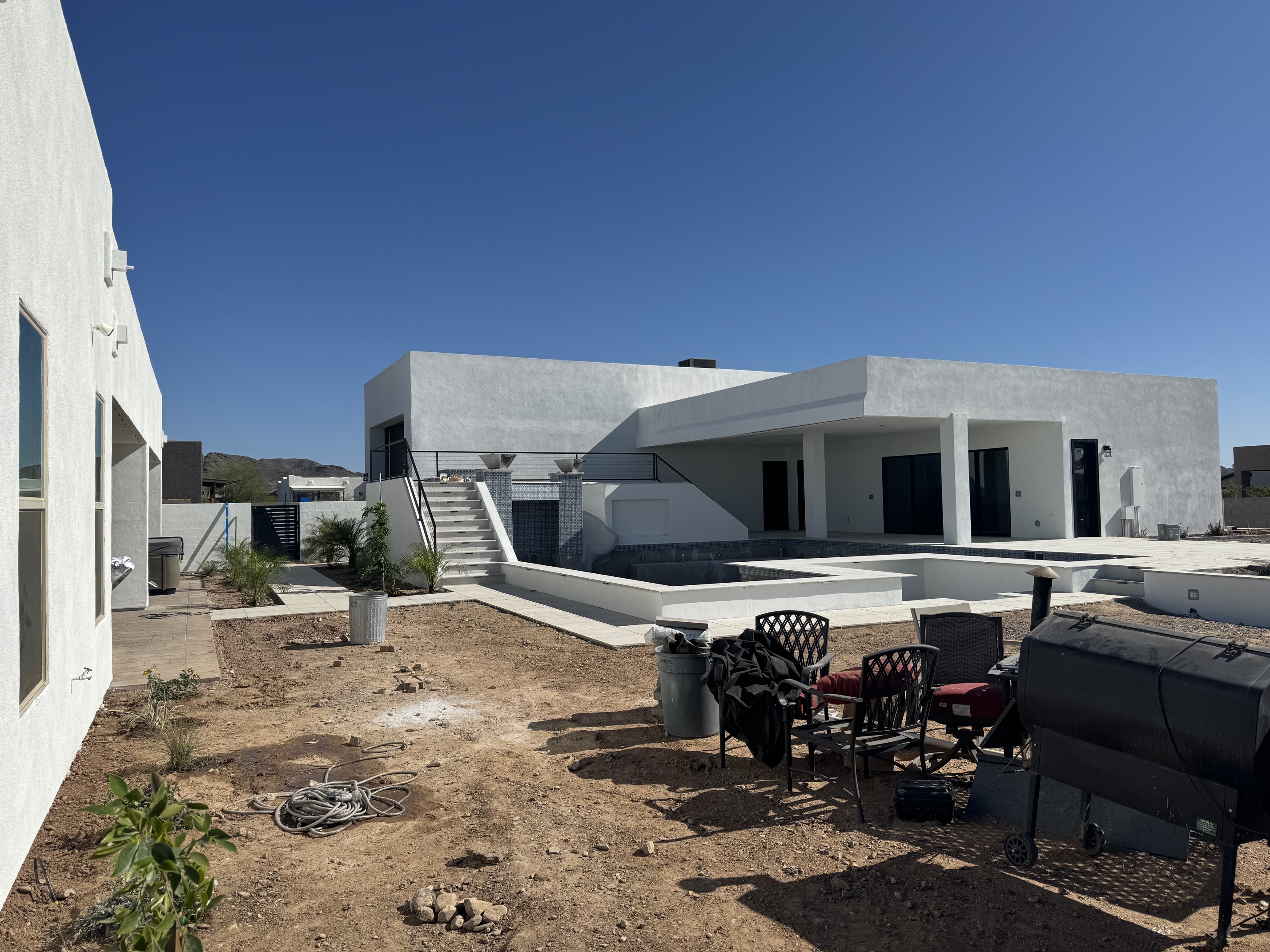 Modern white house with empty pool, patio chairs, barbecue grill, and dirt yard under clear blue sky.