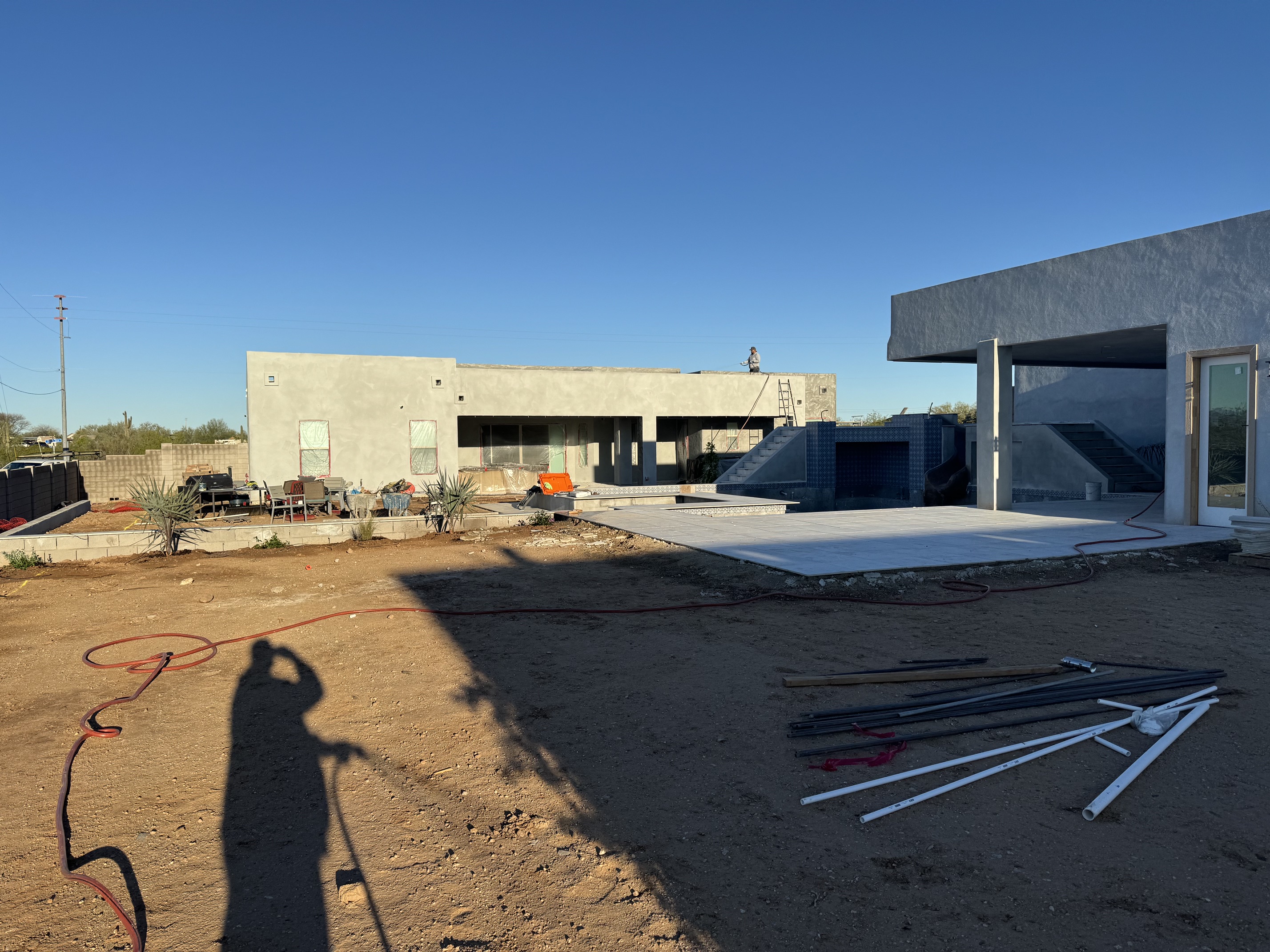 Outdoor construction site with unfinished modern house, patio, and scattered pipes and materials on dirt ground.