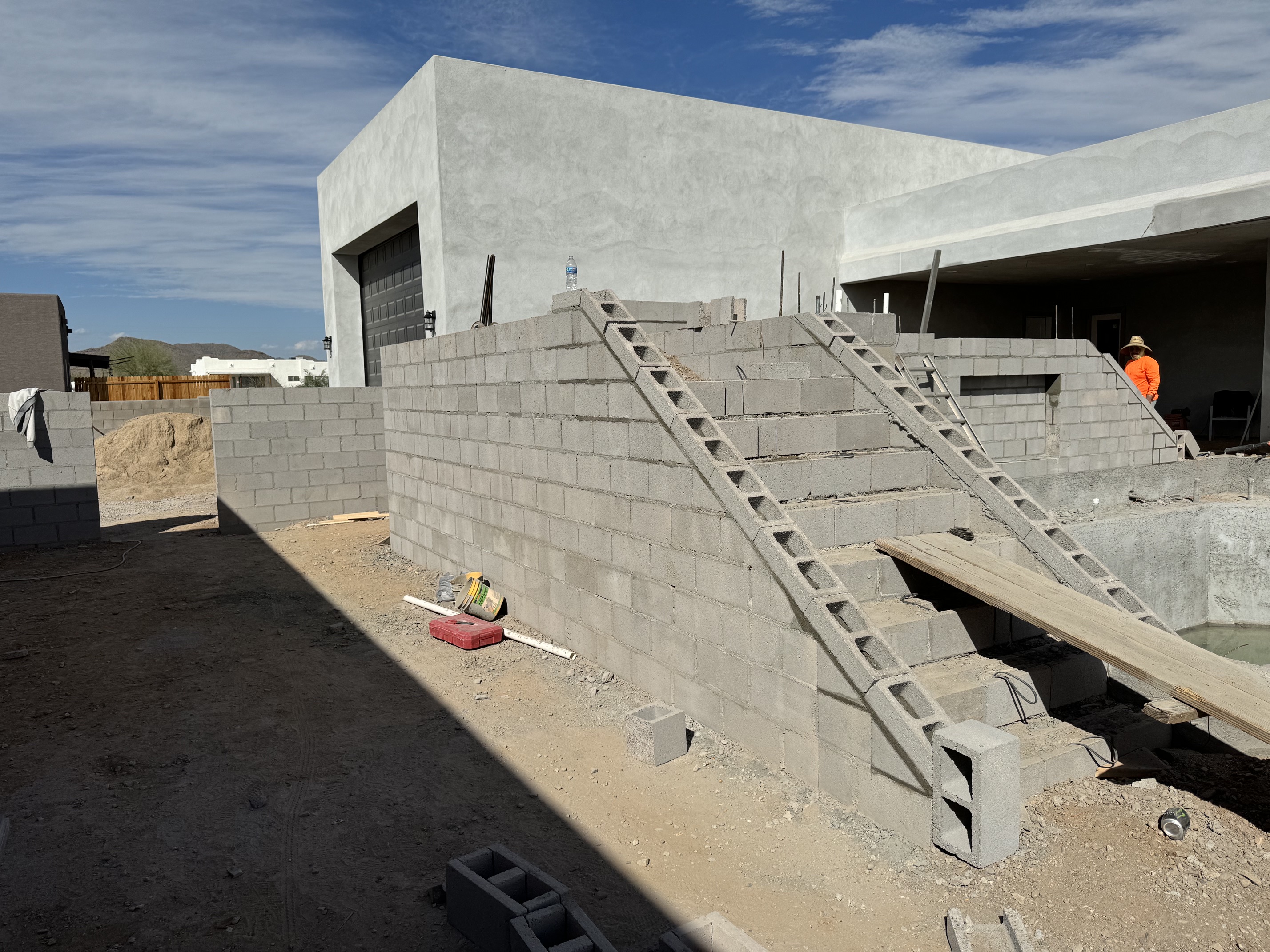 Concrete block walls under construction with stairs and wooden planks in a yard under a blue sky.