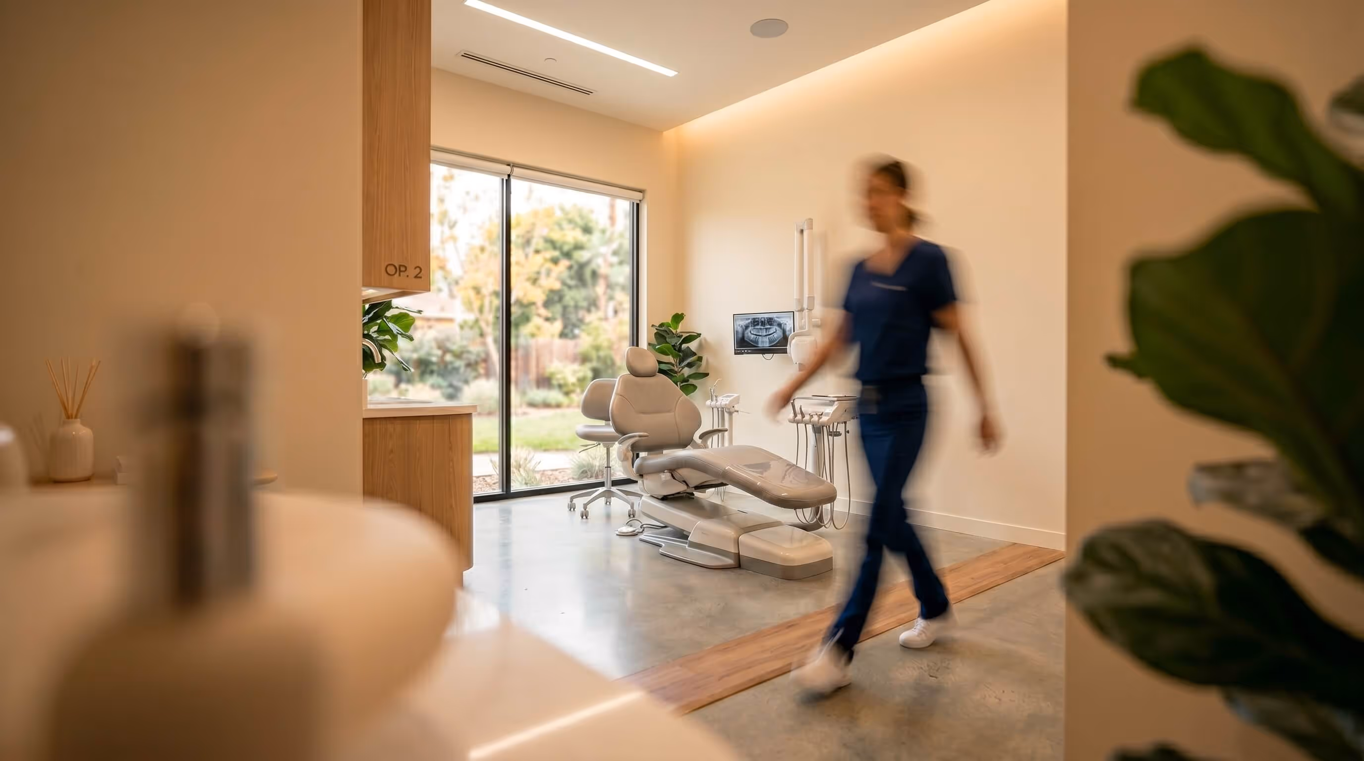 Modern dental office with a dental chair, equipment, a large window showing greenery outside, and a blurred person walking.