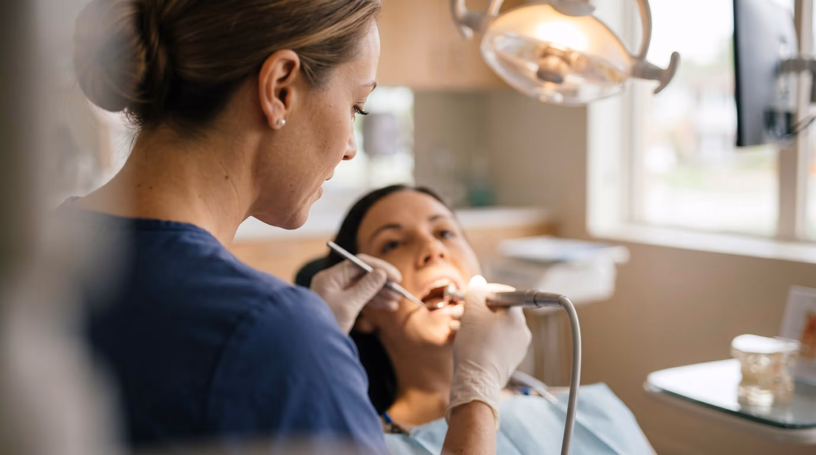 Dentist in blue scrubs examining a patient's teeth with dental tools in a clinic.