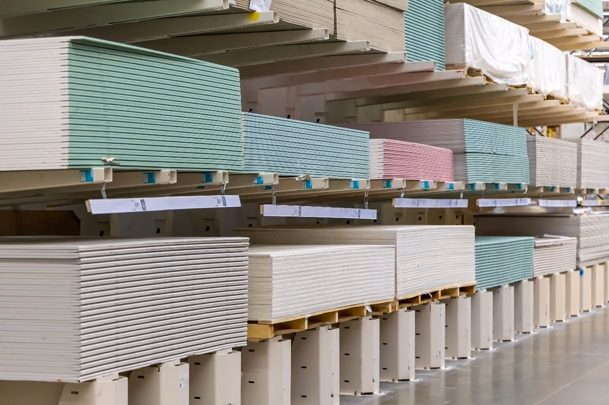 Stacks of plasterboard sheets in different colors on metal shelves in a warehouse aisle.
