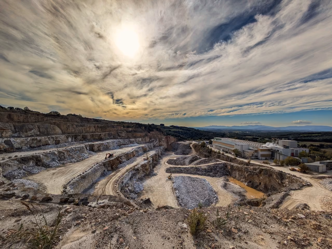 Large terraced quarry with gravel piles and machinery under a cloudy sky with sun.