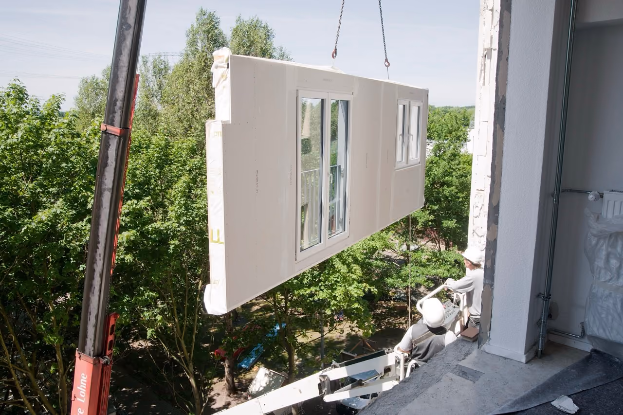 Crane lifting a prefab wall panel with windows to a building where workers guide it inside an open floor.