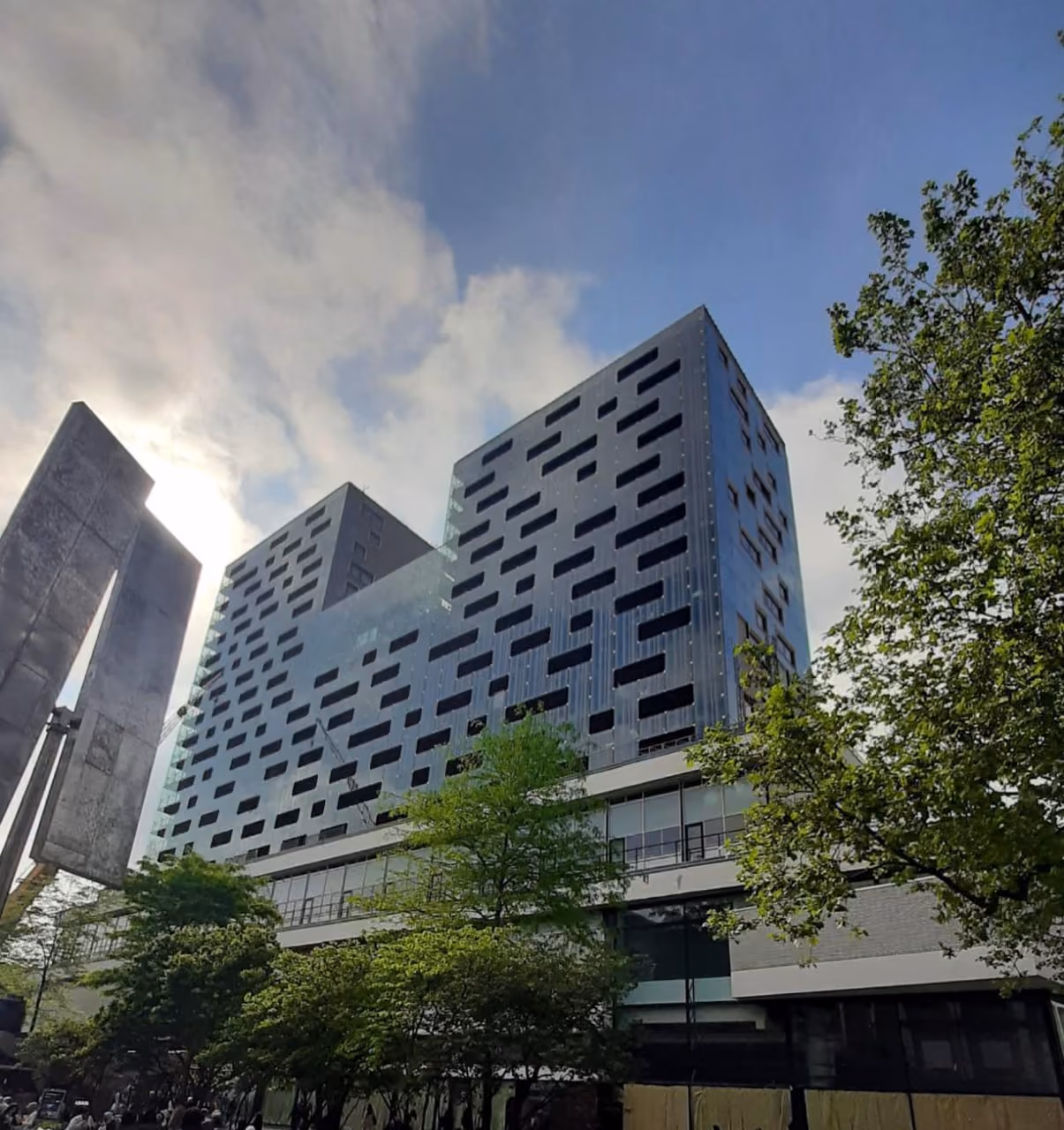 Modern glass building with irregular black window patterns seen behind green trees under a partly cloudy sky.