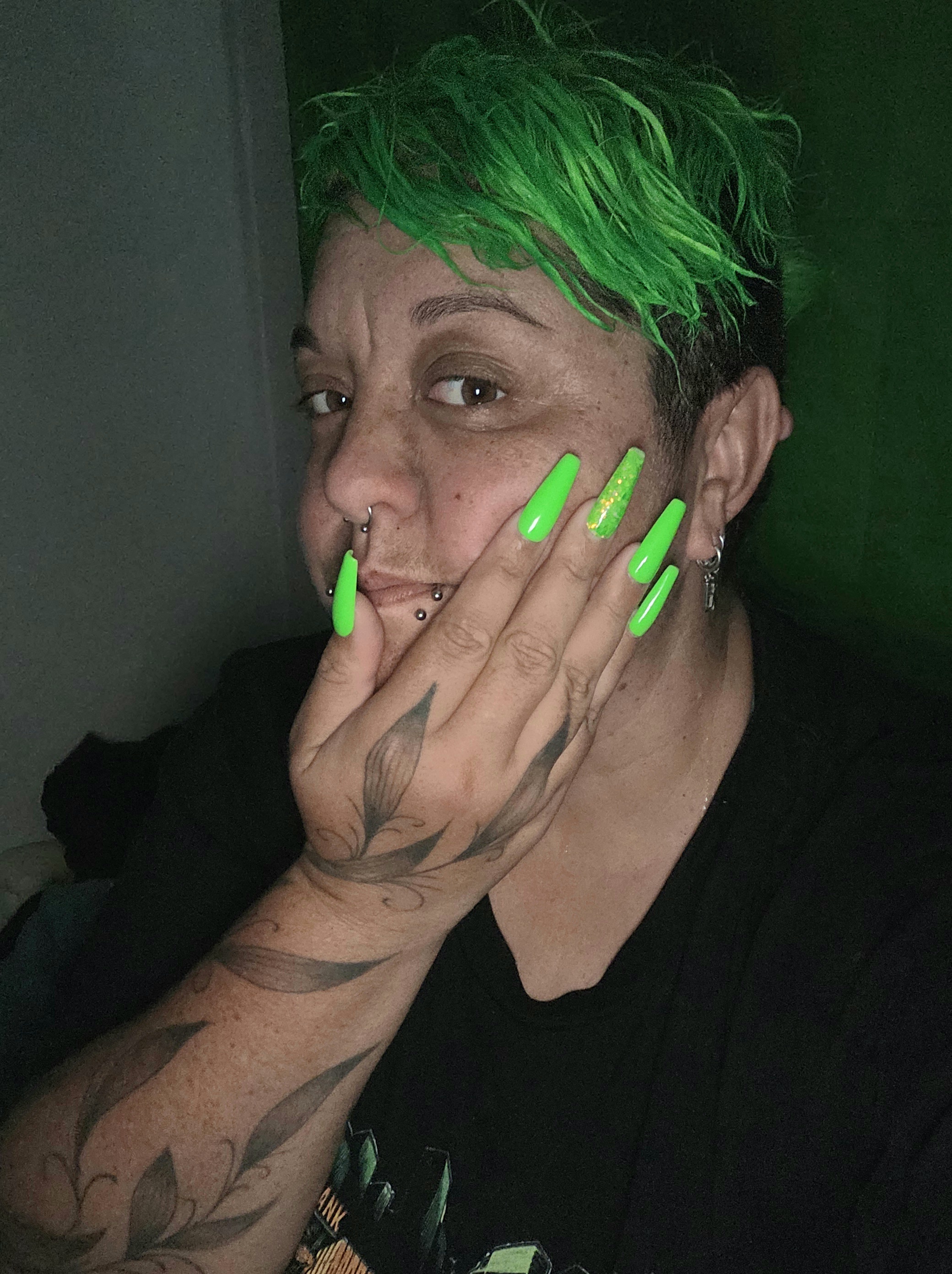 Person with bright green hair and long neon green nails covering part of face, showing tattoos and piercings.