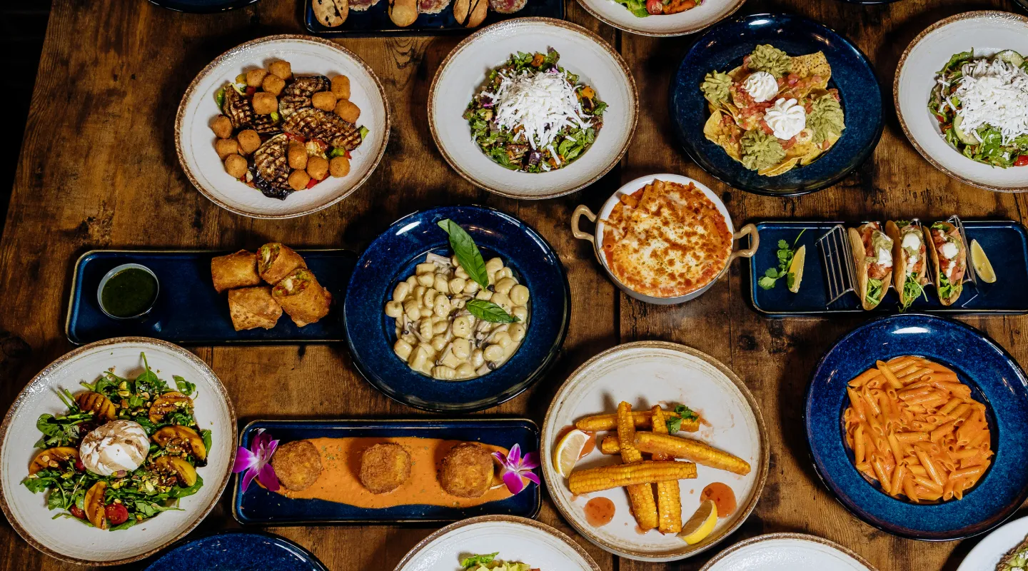 Assorted dishes including salads, pasta, tacos, and fried appetizers arranged on a wooden table.