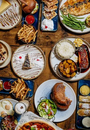 Variety of dishes including burgers, fries, steak, rice, green beans, salad, and desserts on a wooden table.