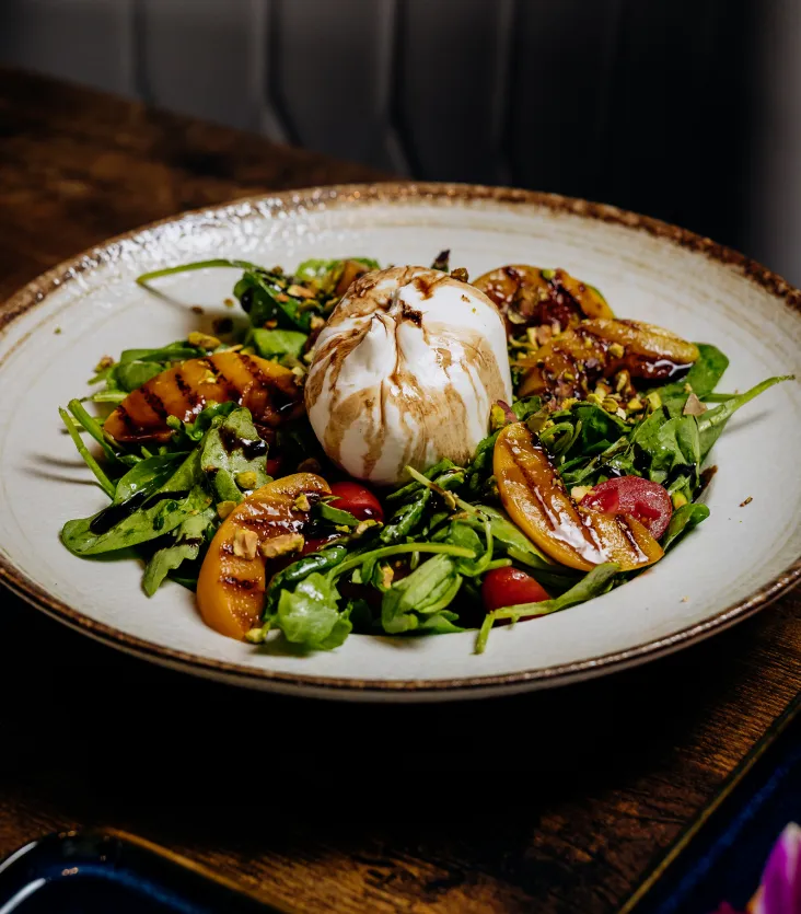 Plate with burrata cheese, grilled peaches, cherry tomatoes, arugula, and balsamic glaze dressing.