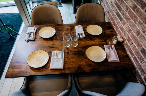 Wooden table set for four with plates, napkins, cutlery, and two empty wine glasses near brick wall.