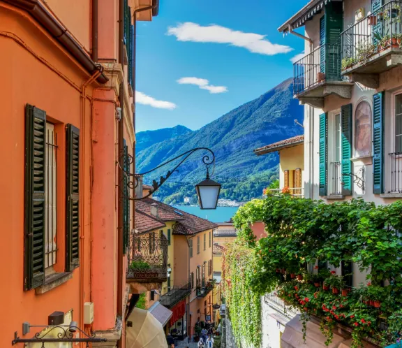 Narrow street with colorful buildings, greenery, and mountain and lake view in the background.