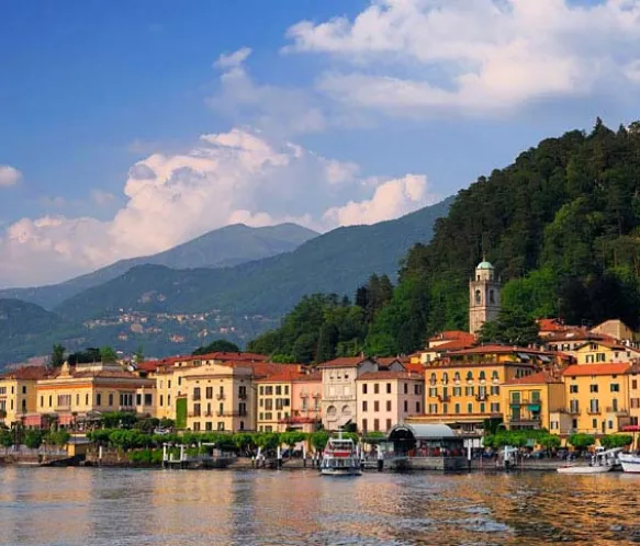 Colorful buildings by a waterfront with boats and a forested hillside under a partly cloudy sky.