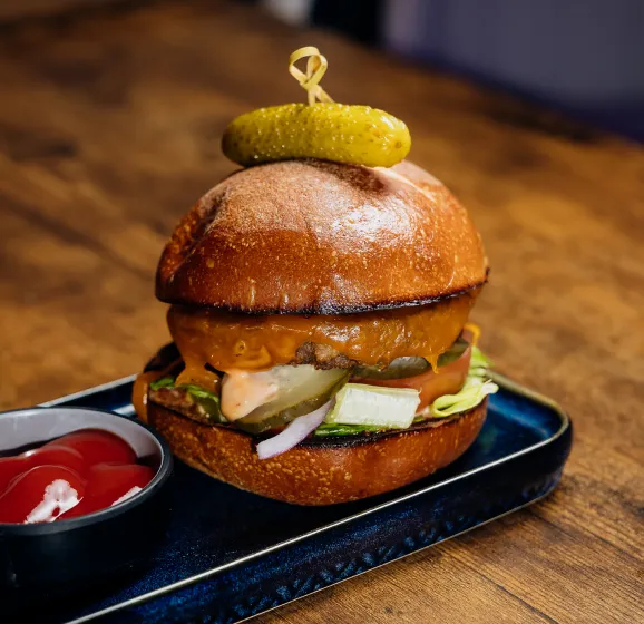 Cheeseburger with lettuce, tomato, pickles, onion, and a pickle on top, served with ketchup.