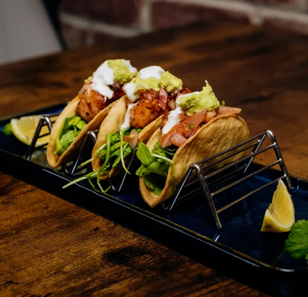 Three tacos with grilled chicken, lettuce, salsa, sour cream, and guacamole on a metal taco holder.
