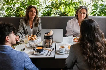 Four people having a business meeting over coffee and food in a bright cafe with plants behind them.
