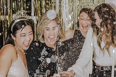 Four women smiling and celebrating with confetti at a festive party with gold streamers in the background.