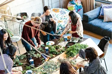 Group of women gathered around a table arranging plants and flowers in a bright room.