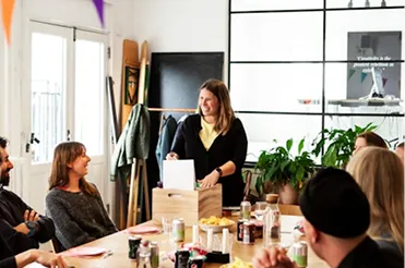 A woman stands and speaks to colleagues around a table in a bright, modern office meeting room.