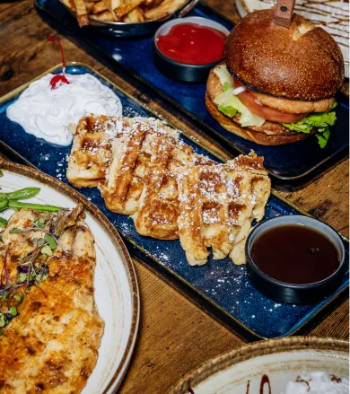 Serving of waffles dusted with powdered sugar, with whipped cream, syrup, and a burger with fries on a wooden table.