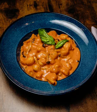 Blue bowl of gnocchi pasta in creamy tomato sauce garnished with fresh basil leaves on wooden table.
