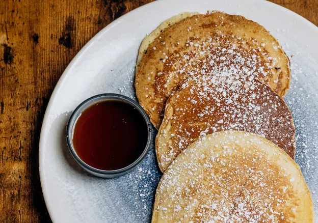 Three stacked pancakes dusted with powdered sugar on white plate with syrup cup on wooden table.