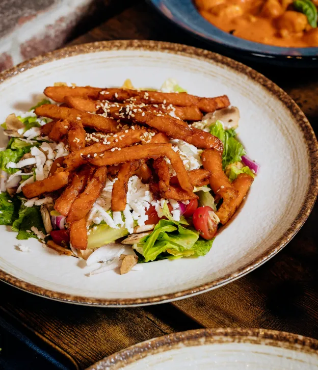Salad with leafy greens, tomatoes, shredded cheese, and sweet potato fries topped with sesame seeds.