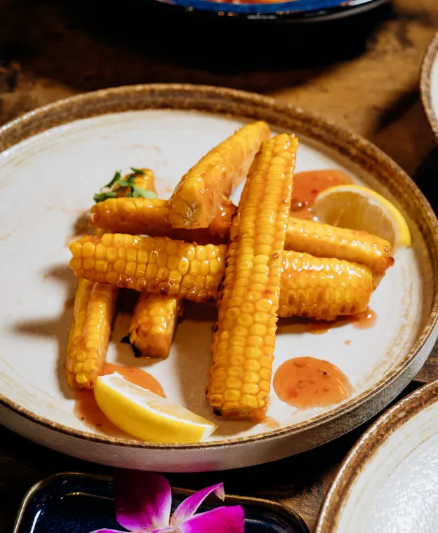 Grilled corn on the cob sticks with sauce and lemon wedges on a ceramic plate.