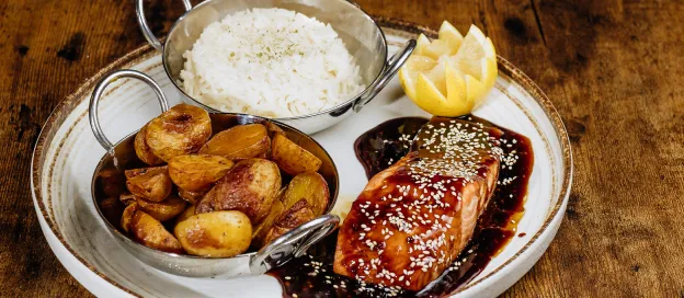 Plate with glazed salmon topped with sesame seeds, roasted potatoes, white rice, and lemon wedge on wooden table.