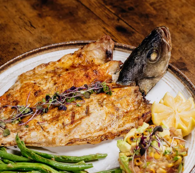 Grilled whole fish with microgreens, lemon wedge, green beans, and corn-avocado salad on a plate.