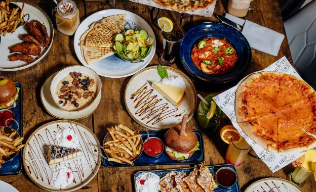 A wooden table filled with various foods including pizza, burger, fries, salad, desserts, and drinks.