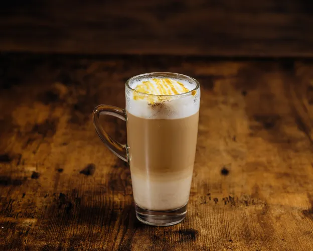 Tall glass mug with layered latte topped with whipped cream and caramel drizzle on a wooden table.