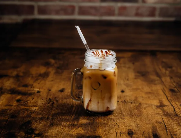 Iced coffee with whipped cream and caramel drizzle served in a mason jar on wooden table.