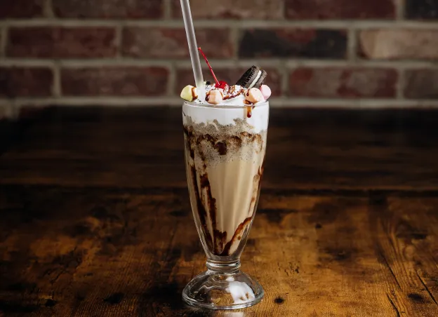Tall glass of chocolate milkshake topped with whipped cream, marshmallows, cherry, and an Oreo cookie.