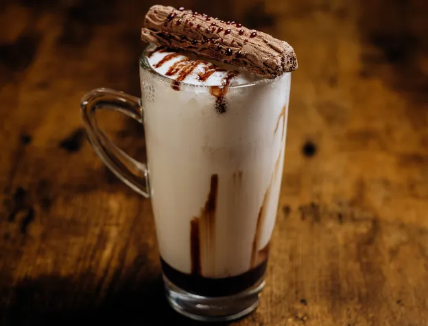 Glass mug of milkshake with chocolate syrup, foam, and a chocolate-covered biscuit on top.