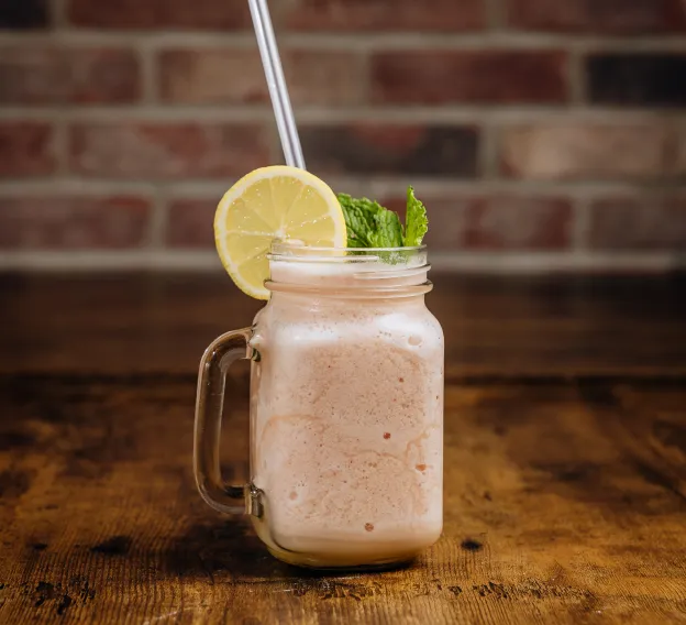 Frosty smoothie in a glass jar with lemon slice, mint, and two straws on wooden table.