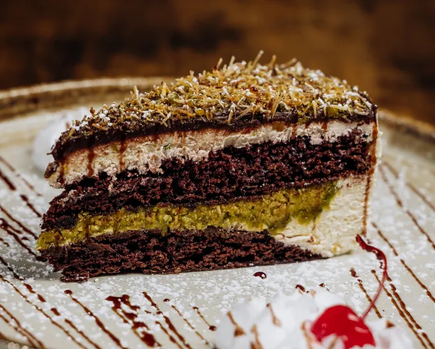 Slice of layered chocolate and pistachio cake with cream on a white plate drizzled with chocolate sauce.