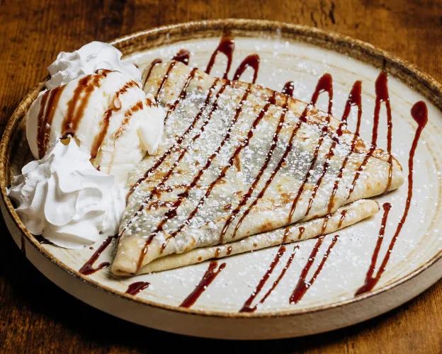 Plate with two folded crepes drizzled with chocolate sauce, powdered sugar, and served with ice cream and whipped cream.