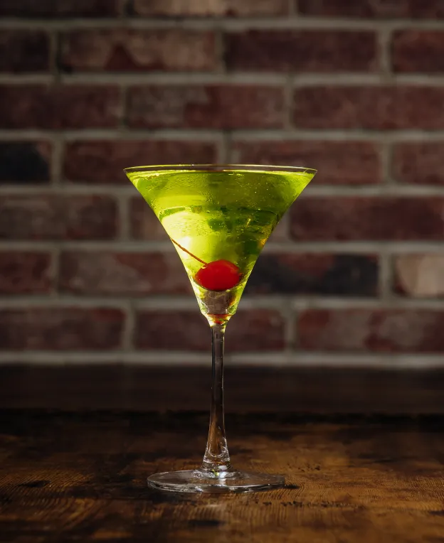 Green cocktail in a martini glass with a red cherry, set against a brick wall background.
