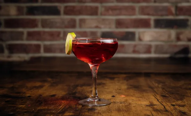 Cocktail glass with a red drink and a lemon slice on the rim on a wooden table with a brick wall background.