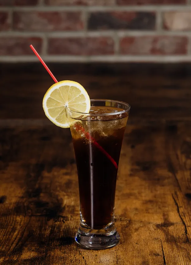Tall glass with iced dark soda, red straw, and lemon slice on rustic wooden table.