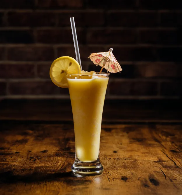 Tall glass of yellow smoothie with a lemon slice, paper umbrella, and clear straw on wooden table.