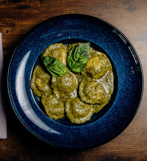 Blue plate with ravioli covered in green pesto sauce, garnished with two fresh basil leaves on wooden table.