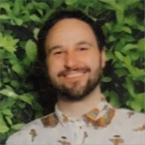 Smiling man with beard and mustache in a patterned shirt in front of leafy greenery.