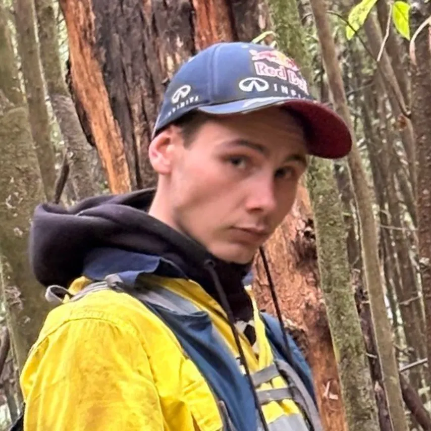 Young man wearing a Red Bull cap and yellow jacket standing in a wooded forest area.