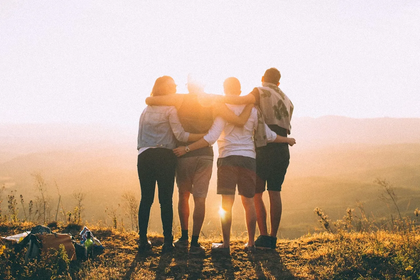 Four friends standing arm in arm outdoors facing a sunset with bags on the ground nearby.