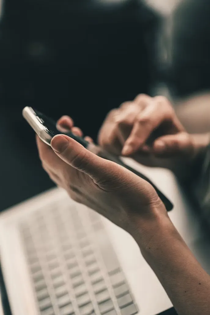 Close-up of hands holding and typing on a smartphone with a laptop keyboard blurred in the background.