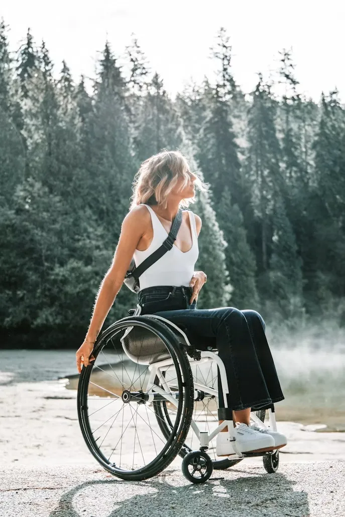 Woman in white top and jeans sitting in a wheelchair outdoors near a misty forest lake.