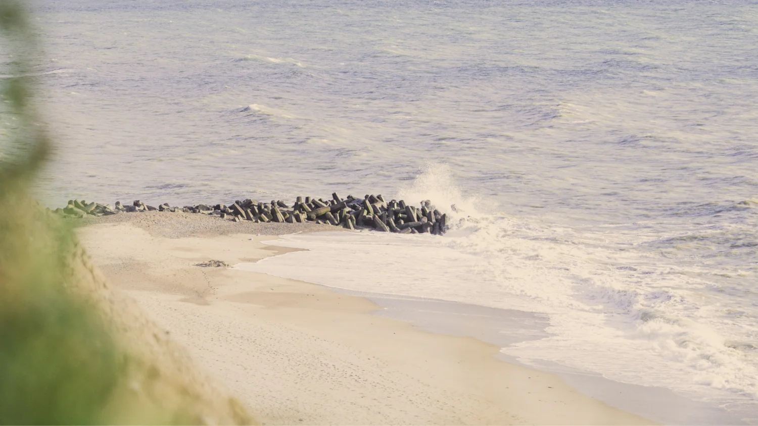 Waves crashing against a cluster of concrete sea defenses on a sandy beach with calm ocean waters.