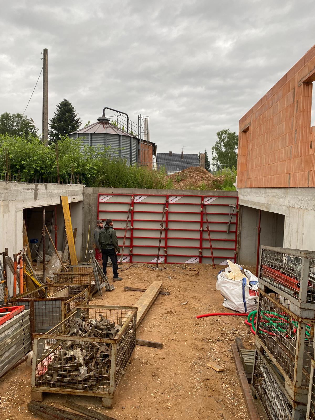 Deux ouvriers sur un chantier avec matériel en métal et murs en béton partiels sous ciel nuageux.
