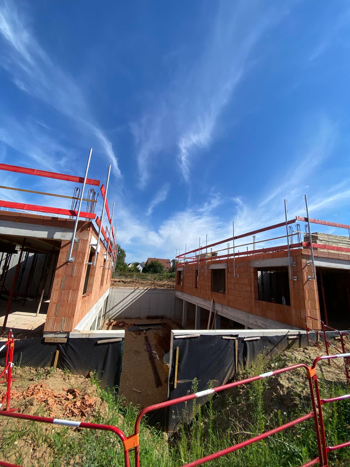 Chantier de maison avec murs en briques rouges et garde-corps rouges sous ciel bleu avec nuages.